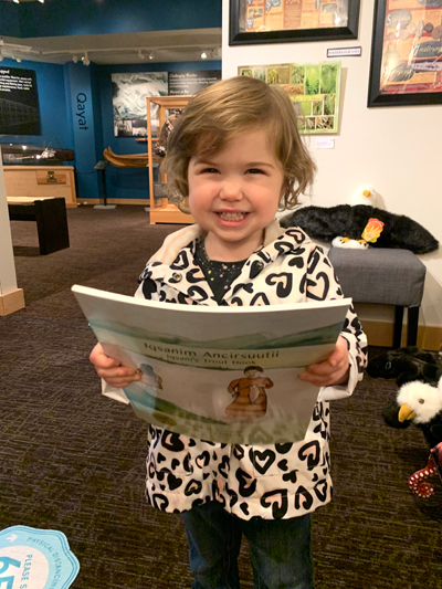 A little girl holds a book while smiling at the camera.